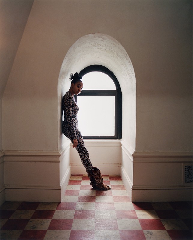 Elegant woman in leopard-print catsuit and boots leans against an arched window in a room with red and white checkered tile flooring.