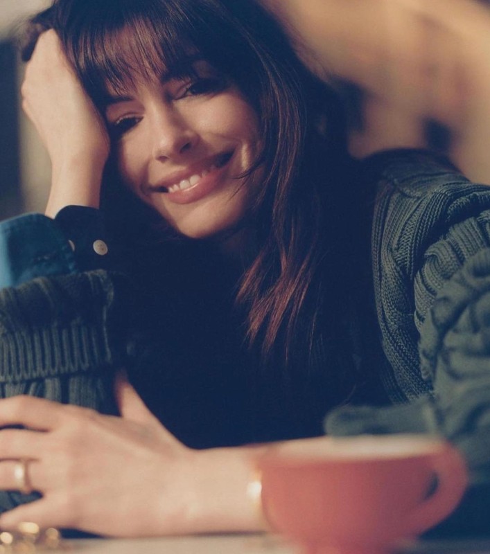 Portrait of a smiling brunette woman in a green sweater leaning on her arms at a table with a pink mug, shot indoors.