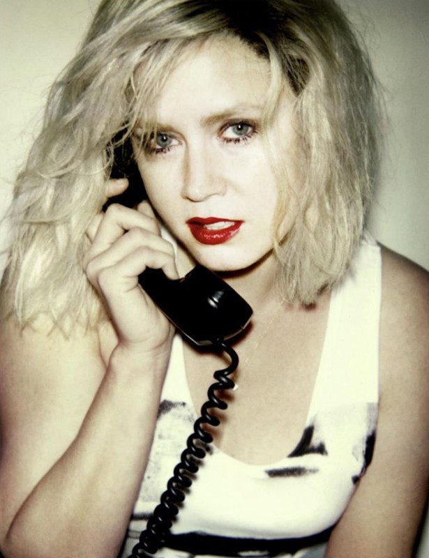 Woman with blonde hair uses a black vintage telephone with a curly cord against a plain backdrop wearing a white tank top and red lipstick.