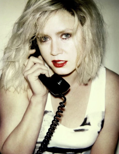 Woman with blonde hair uses a black vintage telephone with a curly cord against a plain backdrop wearing a white tank top and red lipstick.