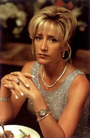 Woman with blond updo and formal attire, silver sequin top, necklace, bracelets, and wristwatch sitting at a table inside.