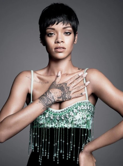 Close-up portrait of Rihanna with short dark hair, wearing a beaded black and teal dress, against a gray background in a photo studio.