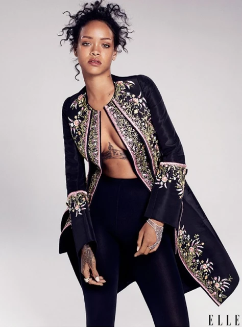 Portrait of woman, Rihanna, posing in a black embellished jacket, leggings, and jewelry against a light gray backdrop in studio lighting.