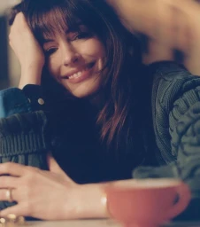 Smiling woman with bangs resting hand on head wearing green knit sweater next to a red cup