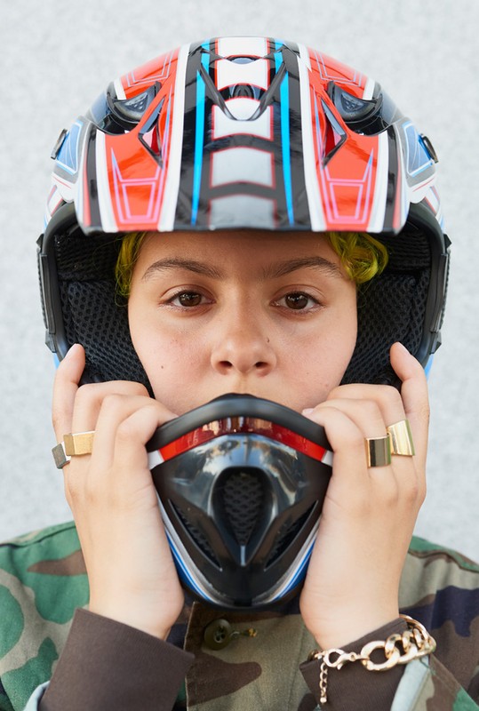 Young woman in a motocross helmet with a camo jacket poses in a close up shot with a neutral expression.