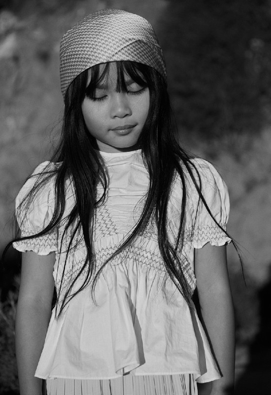 A young Asian girl with eyes closed wears a patterned head covering and a white smocked dress in a black and white portrait.