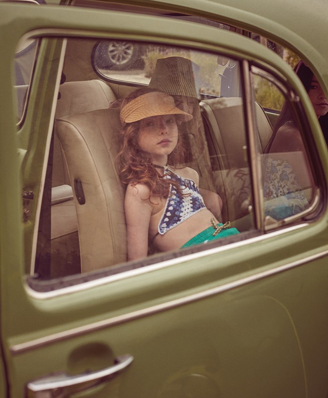 A young girl with red hair and a sun visor sits in the back seat of a vintage green car with tan leather seats, looking out the window.