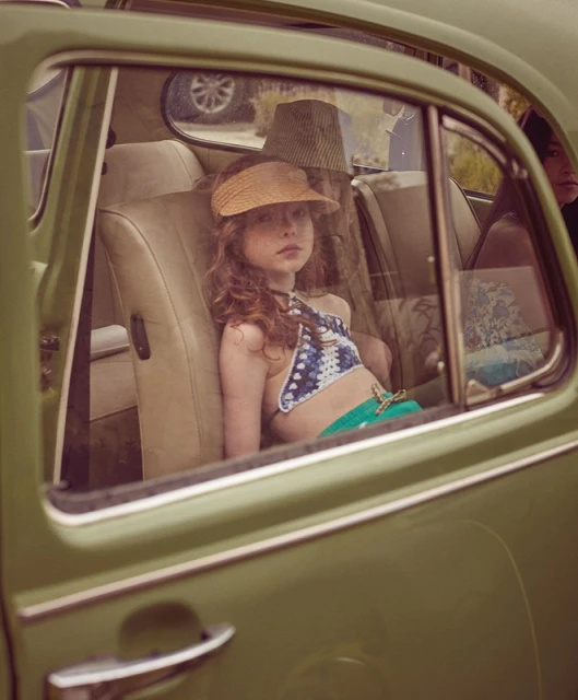 A young girl with red hair and a sun visor sits in the back seat of a vintage green car with tan leather seats, looking out the window.