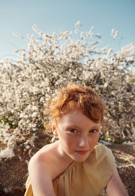Portrait of a red-haired girl with freckles in front of a blooming tree and blue sky, captured in natural light, medium framing.