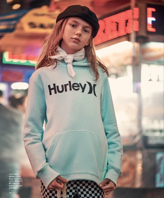 Teen girl in blue Hurley hoodie, black hat, and checkerboard pants poses in front of blurry lights in an urban setting.