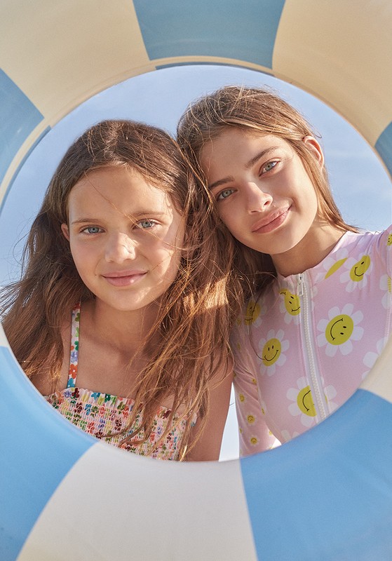 Two young girls smile through a blue-and-white striped life preserver on a sunny day at the beach, wearing colorful swimsuits.