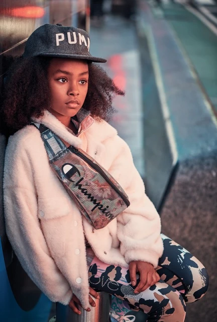 Young child posing in an urban setting wearing a Puma baseball cap, fuzzy jacket, and Champion fanny pack, looking away from camera.