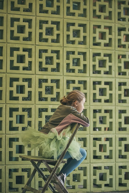 Young girl in tutu sitting on folding chair against geometric patterned wall, casting thoughtful gaze in natural light.