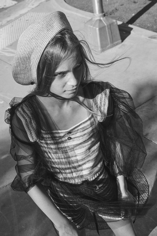 Black and white portrait of a young woman in a plaid top and straw hat, with a concrete surface and pole in the background.