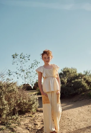 Young red-haired girl in cream-colored outfit standing on a dirt path outdoors surrounded by trees and bushes under a bright blue sky.