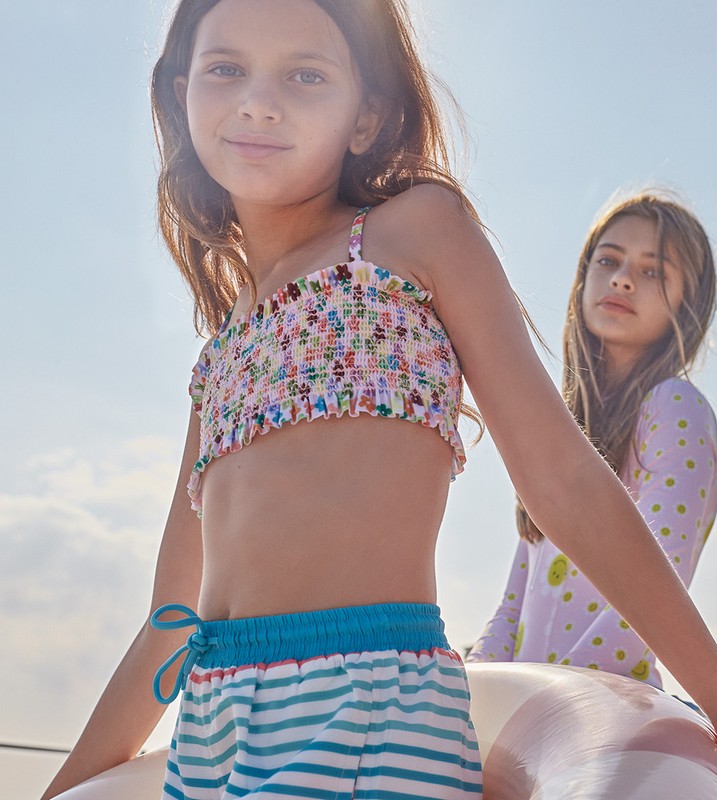 Two young girls pose outdoors. The girl in the foreground wears a floral crop top and striped shorts. The other girl stands behind her.