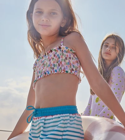 Two young girls pose outdoors. The girl in the foreground wears a floral crop top and striped shorts. The other girl stands behind her.