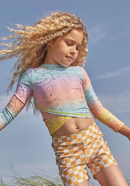 A young girl with blonde curly hair wears a colorful long-sleeved top and checkered shorts outdoors on a sunny day with a blue sky.