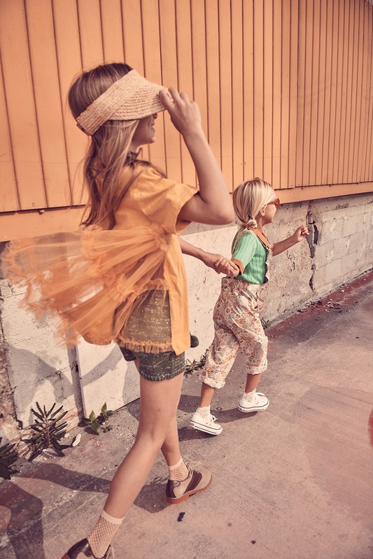 Two children walk together outside in retro-style clothing, one holding the other's hand near a peach-colored wall. 