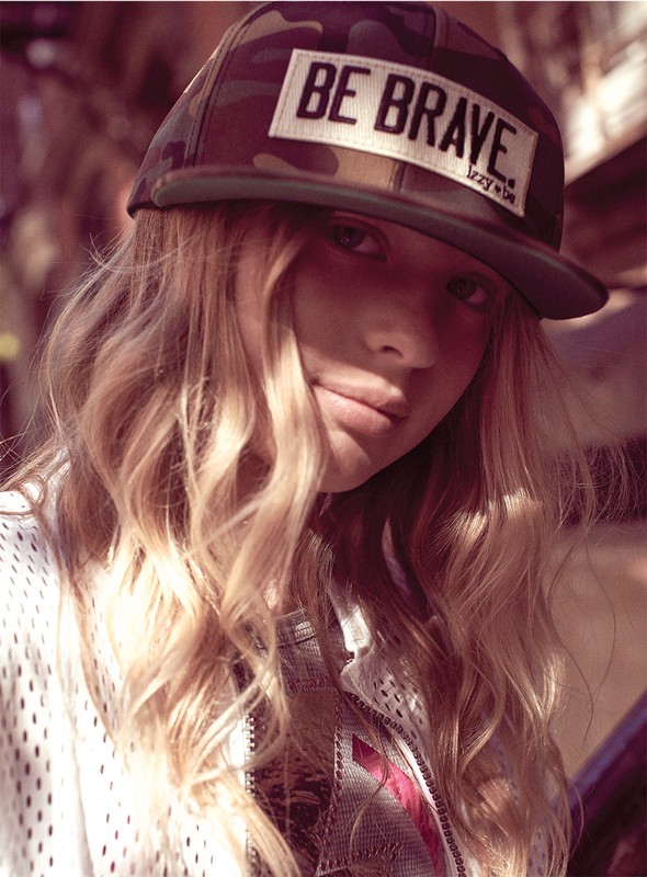 A fair-skinned young woman with blonde hair poses indoors wearing a camouflage baseball cap and a mesh sports jersey.