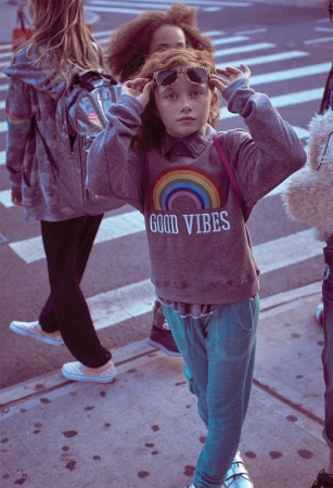 Child wearing a grey sweatshirt with rainbow graphic and adjusting sunglasses on a city street