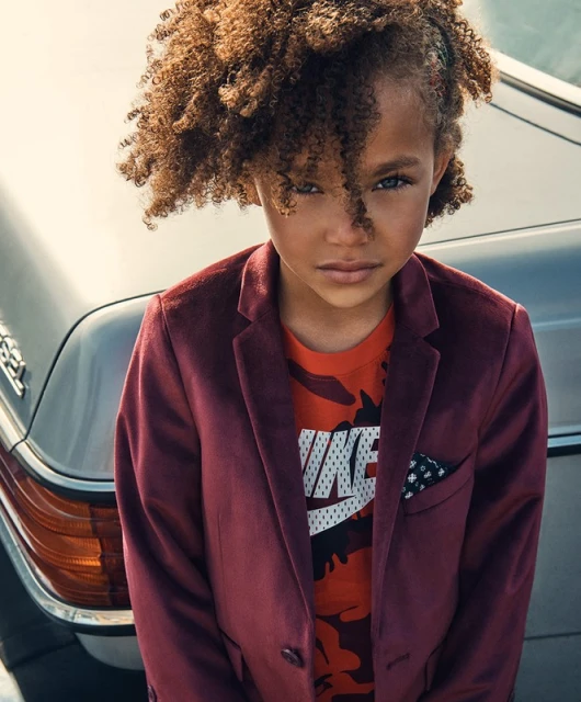 Close-up of stylish child with curly hair wearing a maroon velvet jacket and a graphic t-shirt, posing near a silver car.