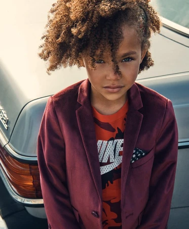 Close-up of stylish child with curly hair wearing a maroon velvet jacket and a graphic t-shirt, posing near a silver car.