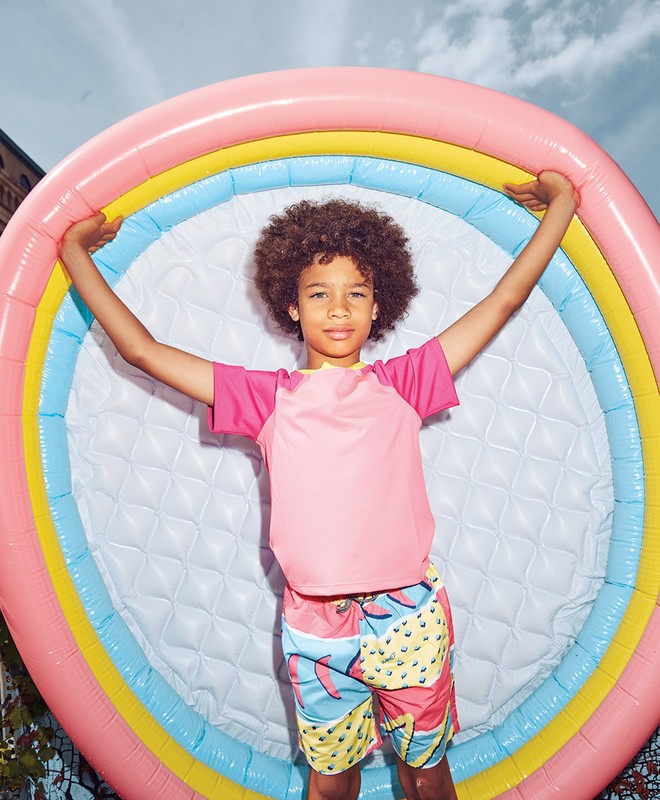 Young boy in swim attire poses in front of a colorful inflatable pool toy, set against an urban background with overcast sky.