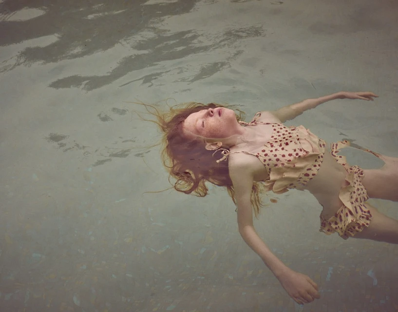 Child in a swimsuit floats on her back in a pool, eyes closed, long hair spread in the water on a bright day with ripples.