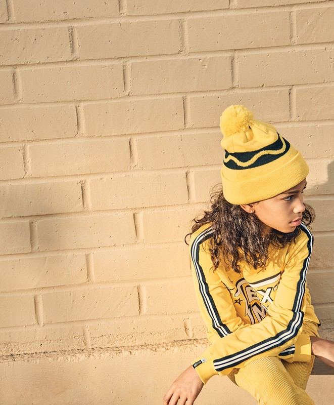 Young child in yellow knitwear: hat with pompom, long-sleeve top with stripes, and matching pants, sitting against a brick wall.