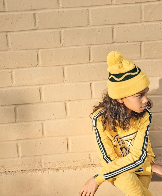 Young child in yellow knitwear: hat with pompom, long-sleeve top with stripes, and matching pants, sitting against a brick wall.