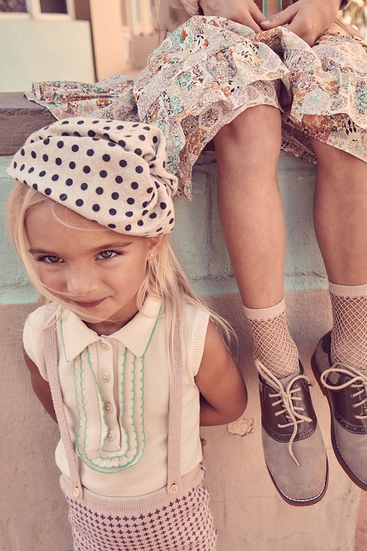 Close-up of a young girl in a polka-dot hat and vintage-style outfit sitting near another person in a floral dress and saddle shoes.