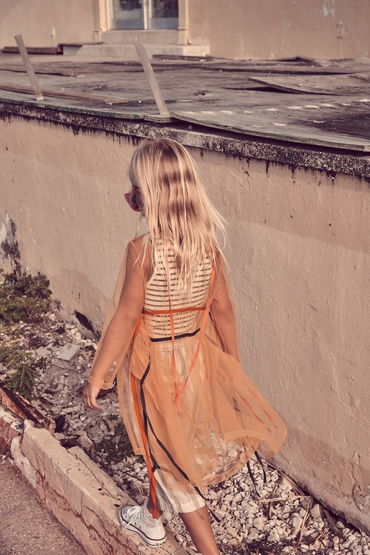 A young girl with long blonde hair wearing a sheer orange dress and white sneakers walks near a decaying wall in an outdoor setting.