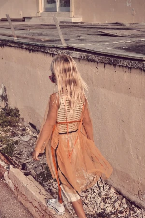 A young girl with long blonde hair wearing a sheer orange dress and white sneakers walks near a decaying wall in an outdoor setting.