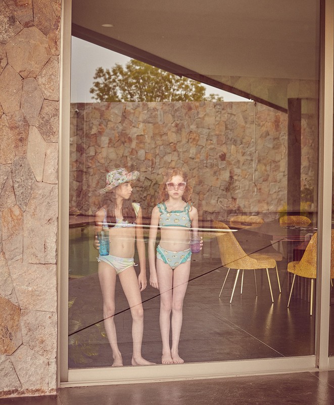 Two young girls in swimwear stand behind a glass wall, reflected in the glass. The background shows a stone wall, pool, and chairs.