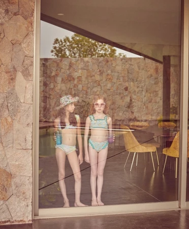 Two young girls in swimwear stand behind a glass wall, reflected in the glass. The background shows a stone wall, pool, and chairs.