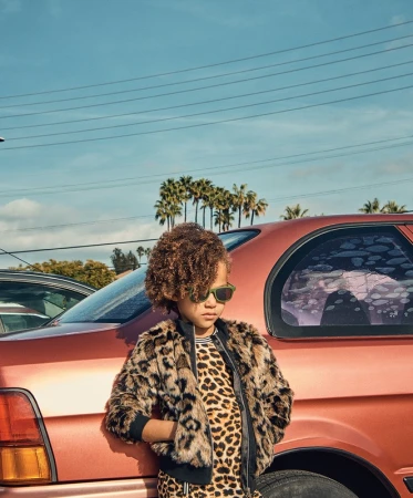 Child with curly hair and sunglasses leaning against a metallic copper car under a clear blue sky