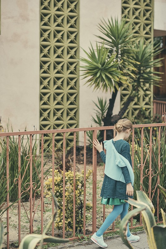 A young girl in a blue dress and tights walks past a pink metal fence, with green plants and architectural details visible in the background.