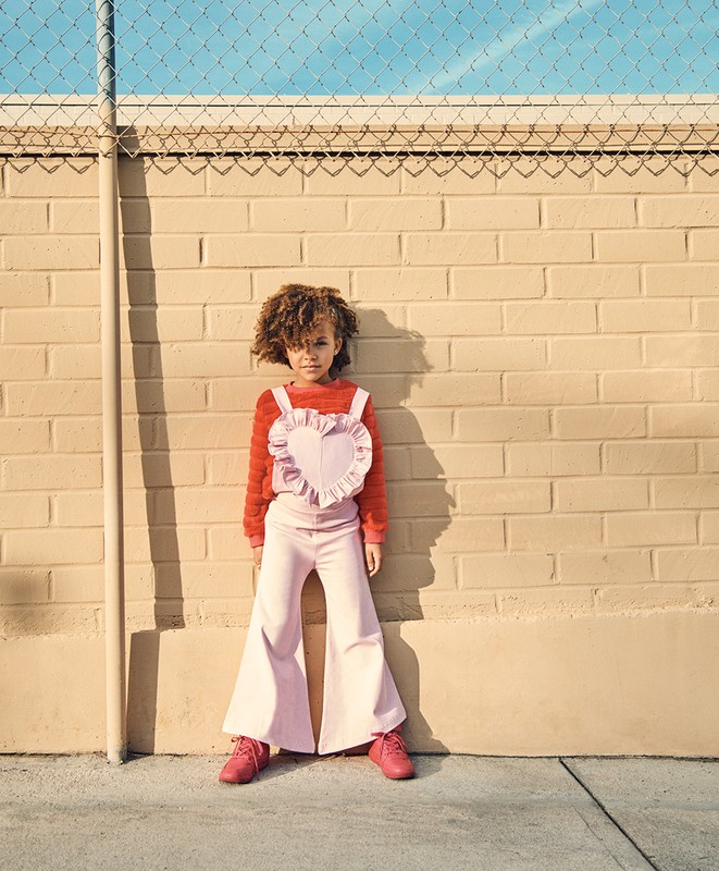 Young girl with curly hair stands in front of a textured brick wall wearing a pink heart top and flared pants in sunlight.