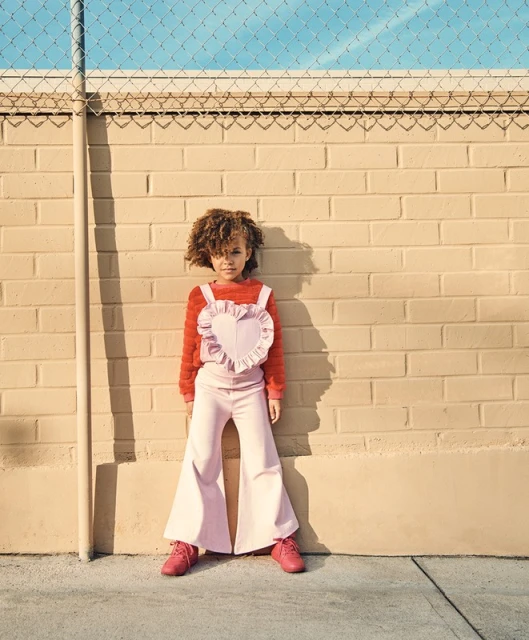 Young girl with curly hair stands in front of a textured brick wall wearing a pink heart top and flared pants in sunlight.
