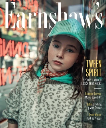 Young girl wearing a textured green and grey sweater, teal cap, and checkered scarf in a street setting