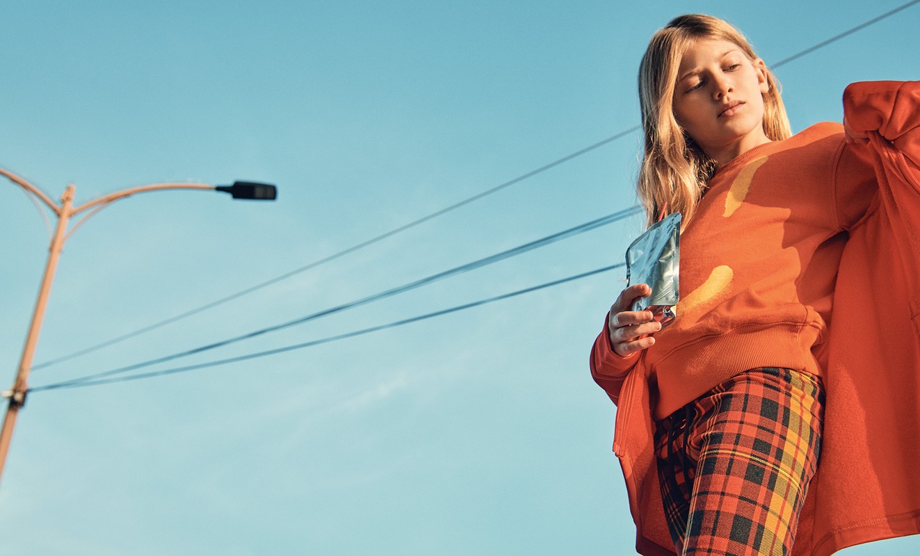 A young fair-skinned woman is shown in an eye-level shot, wearing an orange sweater and plaid pants, set against a bright blue sky.