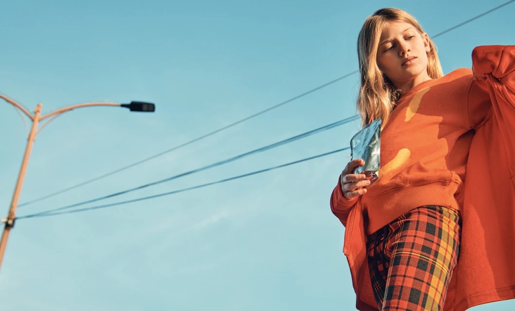 A young fair-skinned woman is shown in an eye-level shot, wearing an orange sweater and plaid pants, set against a bright blue sky.