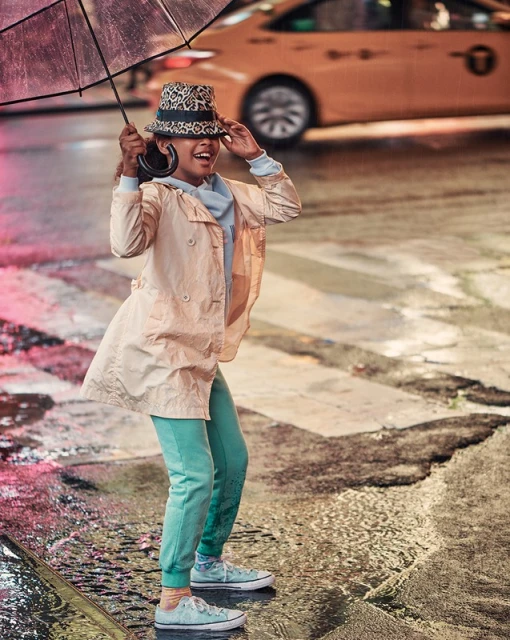 Young girl with an umbrella stands in the rain in a city, wearing a hat, light jacket and teal sweatpants, with taxi in background.