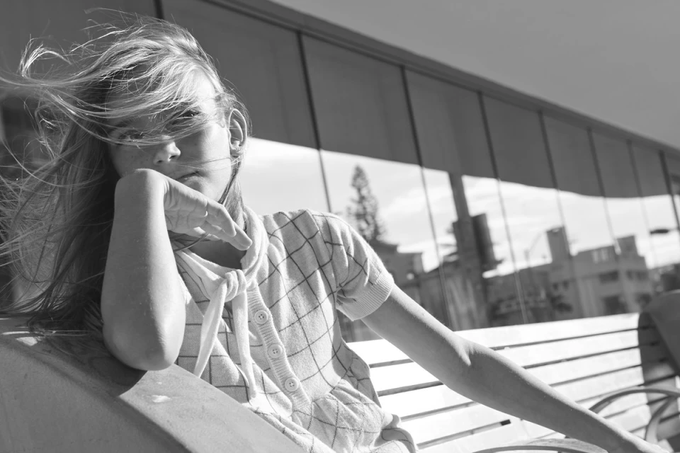 Black and white portrait of a young woman seated on a bench, wearing a patterned sweater, wind blowing through her hair, with a building backdrop.