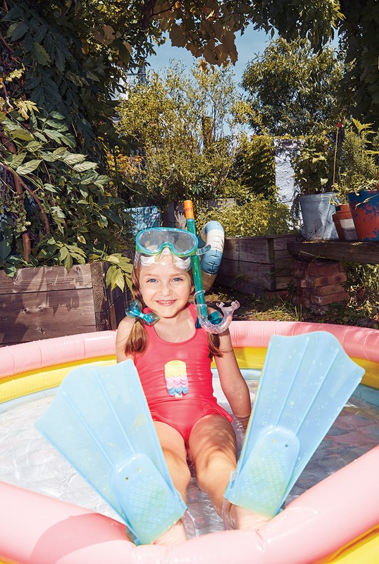 A happy child in a swimsuit and swim gear sits in an inflatable pool outdoors, surrounded by lush green foliage on a sunny day.