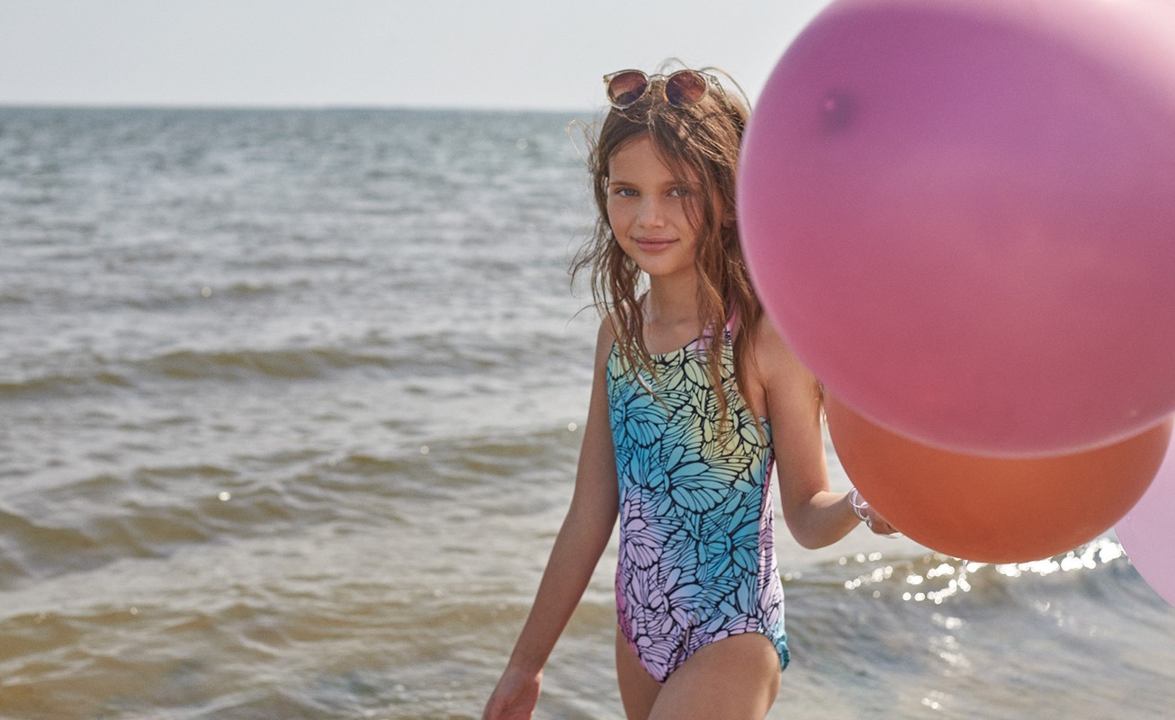 A young girl in a blue gradient swimsuit holding pink and orange balloons while wading in the ocean.