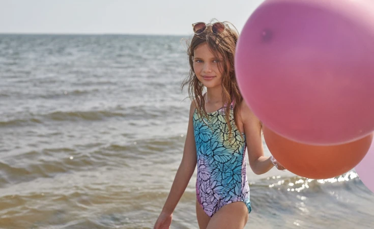 A young girl in a blue gradient swimsuit holding pink and orange balloons while wading in the ocean.