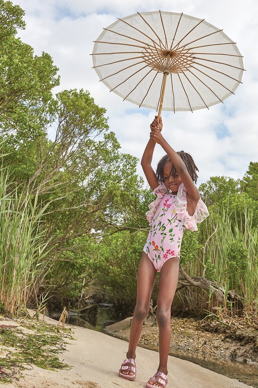 A young African American girl in a floral swimsuit poses on a beach, holding a white parasol overhead, surrounded by greenery and sky.
