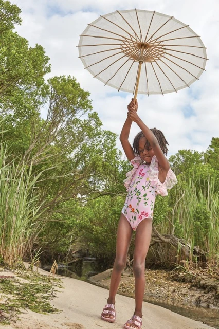 A young African American girl in a floral swimsuit poses on a beach, holding a white parasol overhead, surrounded by greenery and sky.